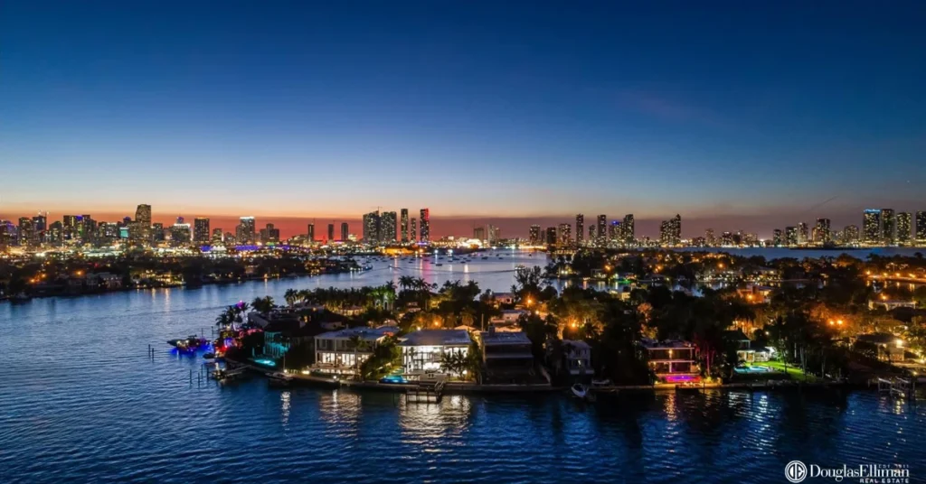 Aerial panoramic photograph of the exclusive Venetian Islands in Miami Beach, showcasing luxurious waterfront homes with private docks during a vibrant sunset over Biscayne Bay. The Miami skyline is visible in the distance.