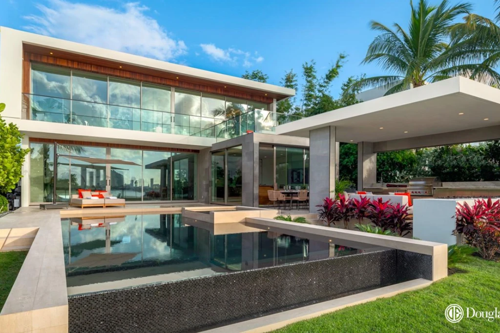 A modern waterfront mansion in Miami Beach at sunset, featuring a glowing infinity pool, a fully equipped outdoor kitchen, and lush tropical landscaping overlooking the bay.