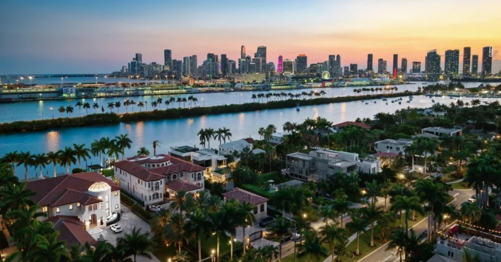 A luxurious two-story Mediterranean-style waterfront mansion in Miami, illuminated at dusk with the glittering city skyline visible across the bay, featuring a large swimming pool and private dock.