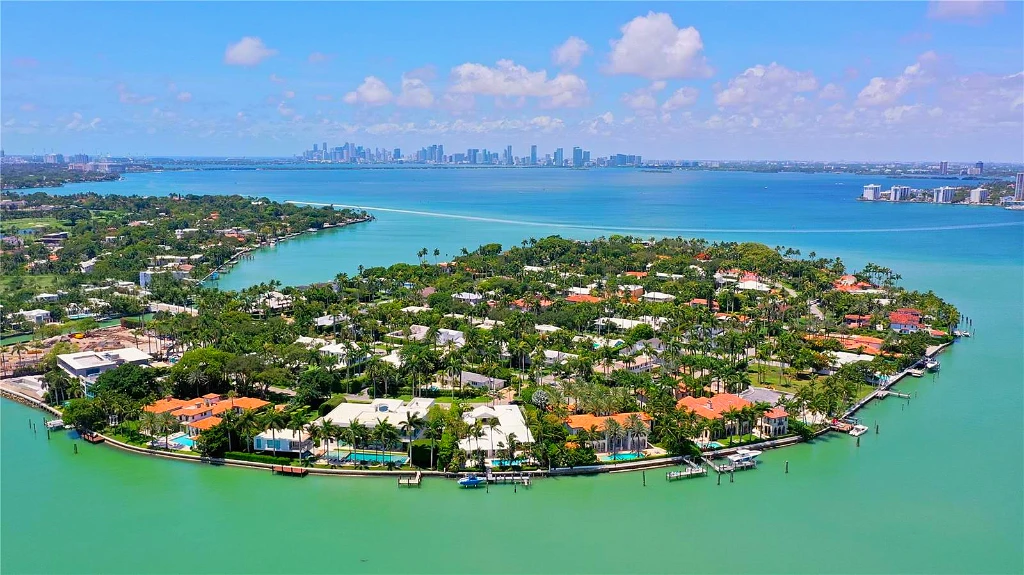 Aerial view of La Gorce Island in Miami Beach, showcasing several luxury waterfront estates with private swimming pools, lush landscaping, and private boat docks lining the turquoise waters of Biscayne Bay.